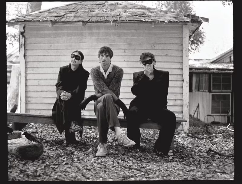Three adults wearing masks sit on a bench in front of a wooden building outdoors on a cloudy day. The scene is in black and white.
