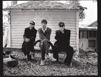 Jacob Elordi seated on a bench outdoors flanked by two figures wearing black eye masks in front of a rustic wooden shed.