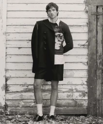 Actor Jacob Elordi wearing a layered dark coat, graphic sweater, shorts, and white socks against a weathered white wooden wall.