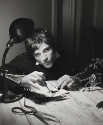 Actor Jacob Elordi leaning over a wooden table working with tweezers and scissors beside a desk lamp in black and white.