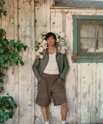 Jacob Elordi wearing an olive green coat draped with white flowers, a white tank top, and brown shorts against a rustic wooden wall.