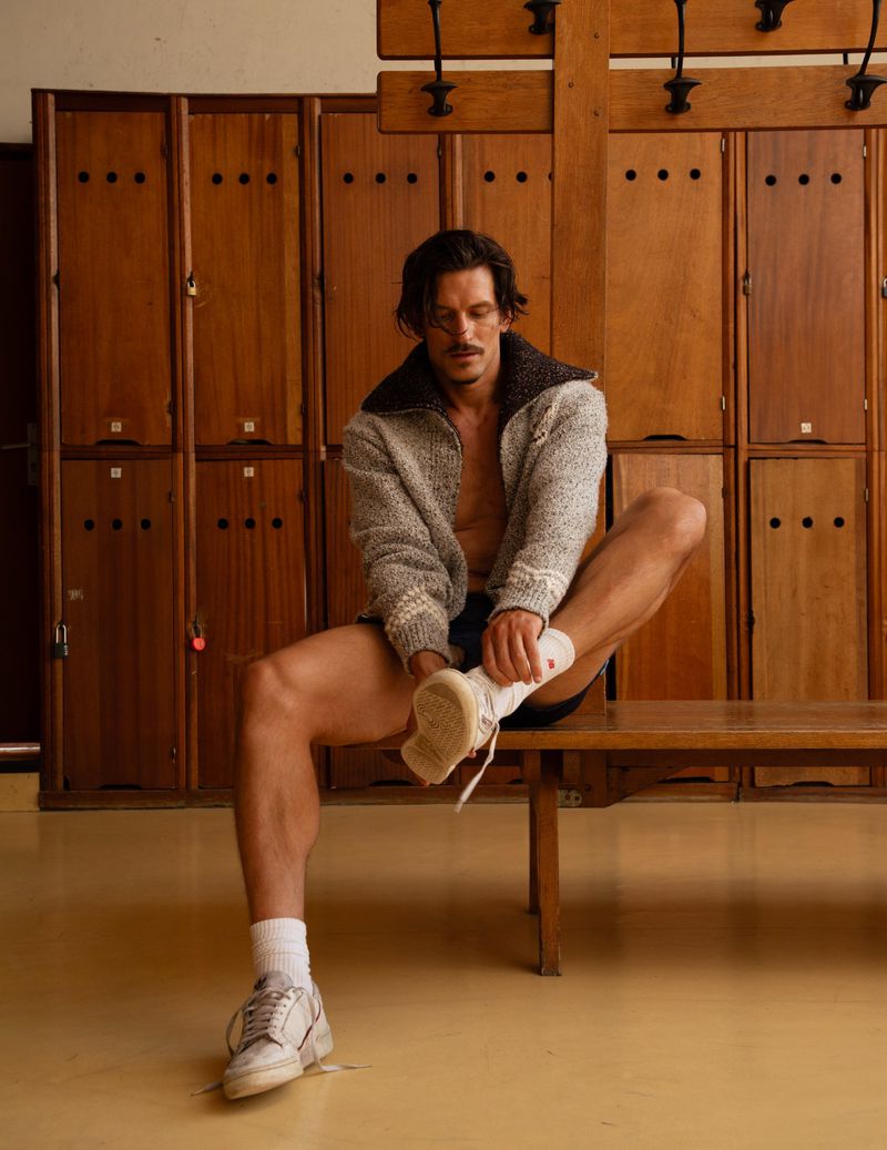 A man sits on a bench in a locker room, putting on a white sneaker. He is wearing a grey sweater and black shorts.