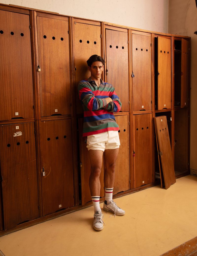 A young adult male poses indoors in a striped rugby shirt, white shorts and sneakers, standing against a backdrop of wooden lockers.