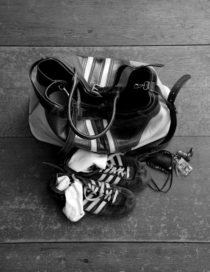 Black and white image featuring a duffel bag with white stripes, a pair of sneakers with socks, and keys on a weathered wooden surface.