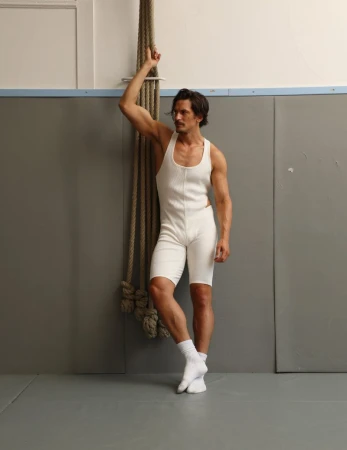 Muscular adult male in a wrestling singlet and white socks poses in an indoor setting, leaning against a climbing rope near a gray wall.