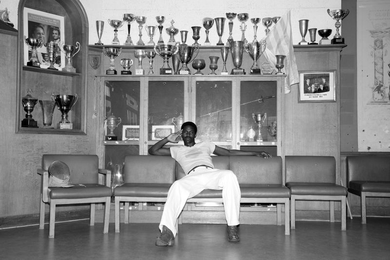 Monochrome portrait of a man sitting on a couch in a room filled with trophies and memorabilia on shelves and cabinets. 
