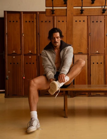 Male model seated on a wooden bench adjusting his sneaker in a locker room for Harper's Bazaar France Homme.