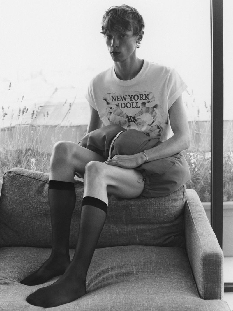 Black and white portrait of a young man with pale skin sitting on a couch in sheer socks and shorts with a t-shirt that says New York Doll.
