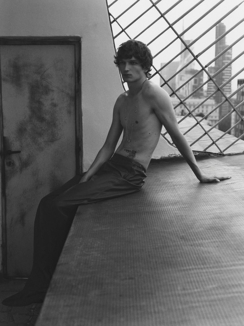 Black and white portrait of a shirtless man sitting on a ledge in front of a window with a city skyline in the background.