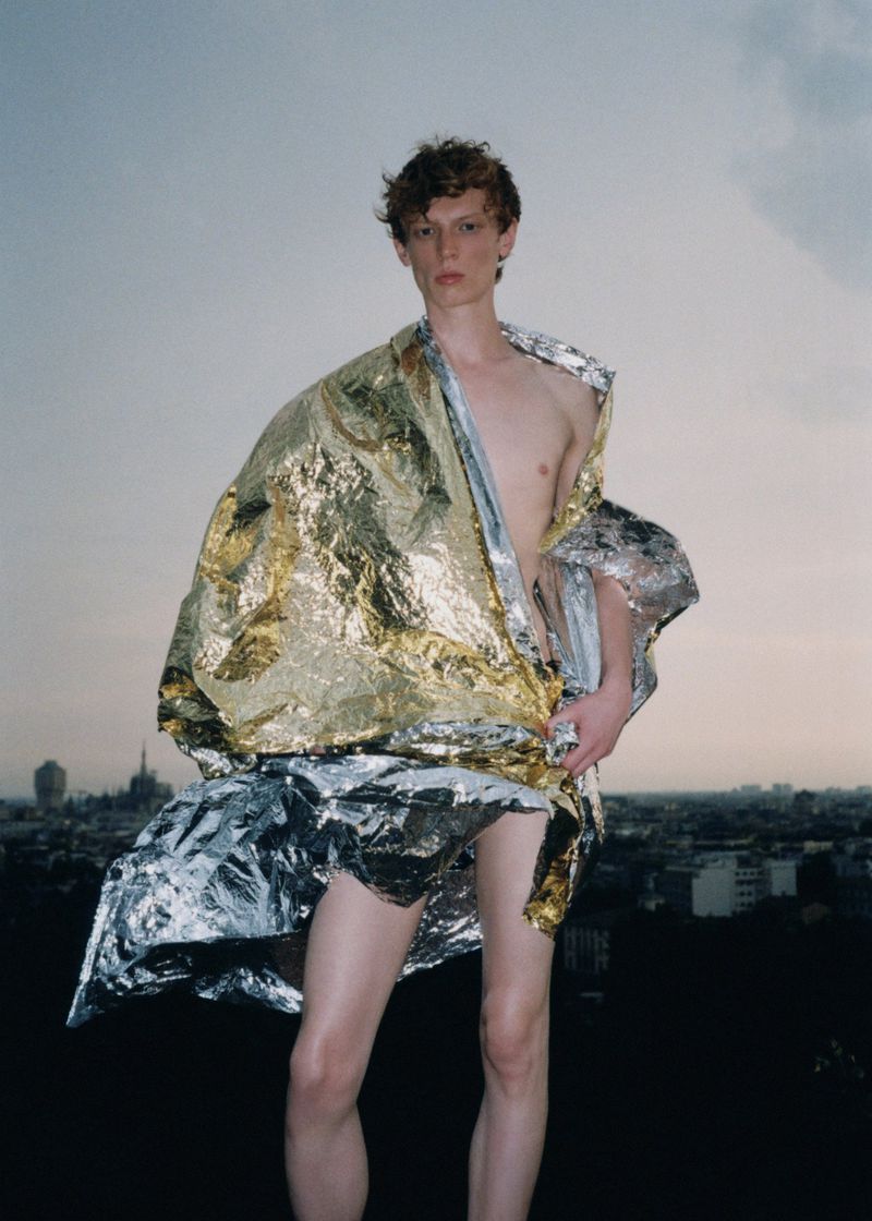 Adult male model draped in a shiny gold and silver blanket poses outdoors on a rooftop with a city skyline in the distance.