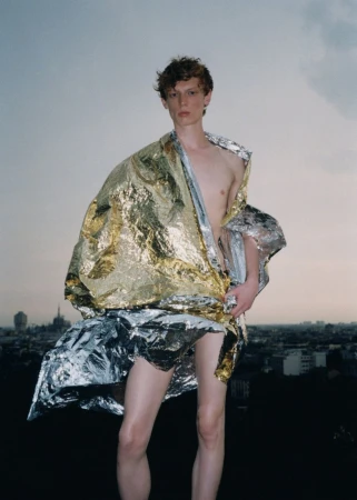 Adult male model draped in a shiny gold and silver blanket poses outdoors on a rooftop with a city skyline in the distance.