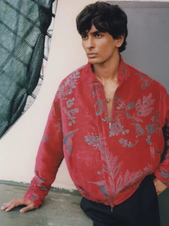 Model wearing a red floral patterned zip-up jacket and black trousers posing outdoors while leaning against a ledge.