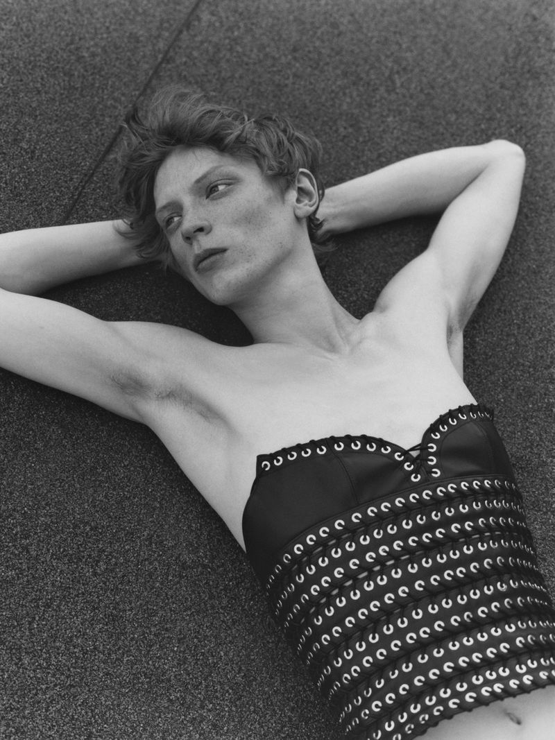 Monochrome portrait of a person lying down, wearing a top with grommet details, against a neutral background, showcasing fashion.