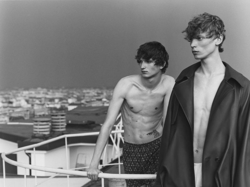 Two young adult men pose on a deck overlooking an urban landscape under a cloudy sky in a black and white fashion editorial.
