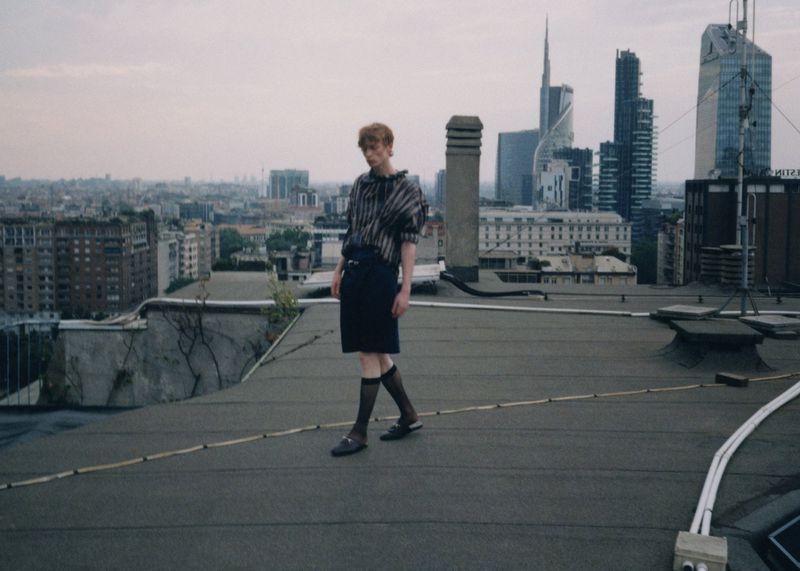 A fair-skinned adult stands on a rooftop wearing dark clothing, hosiery, and loafers in an urban setting with skyscrapers behind them.