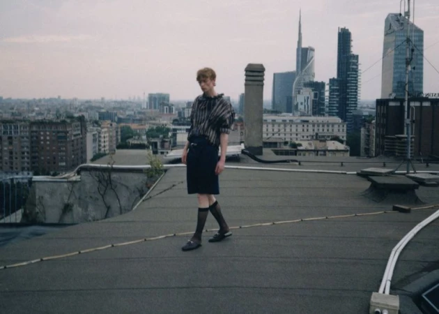 A fair-skinned adult stands on a rooftop wearing dark clothing, hosiery, and loafers in an urban setting with skyscrapers behind them.