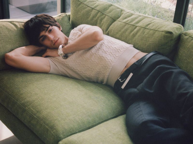 A young man reclines on a green sofa wearing a cream textured knit top, dark trousers, and a chunky silver bracelet.