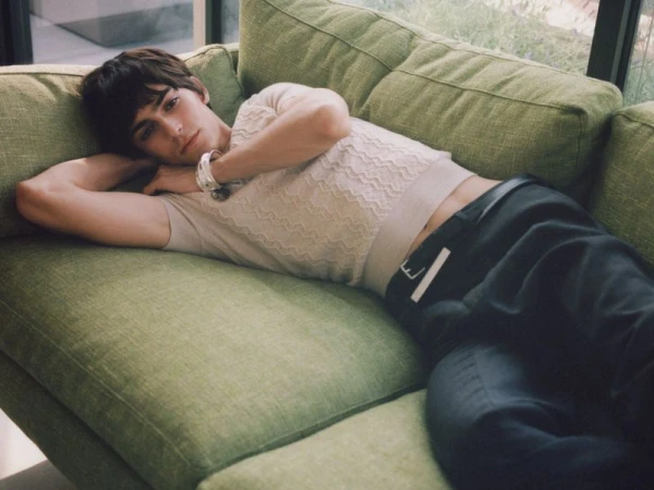 A young man reclines on a green sofa wearing a cream textured knit top, dark trousers, and a chunky silver bracelet.