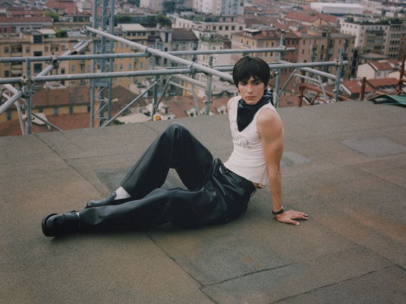 Adult model poses on a rooftop in a white top and black trousers, with a cityscape backdrop during daytime, creating an editorial fashion image.
