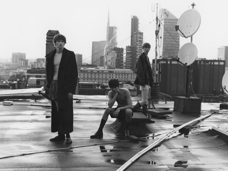 Three adult males pose on a rooftop with the city skyline in the background on an overcast day in black and white.