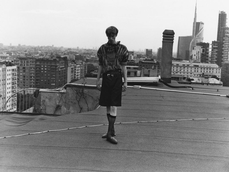 Monochrome fashion shot of a young adult standing on a rooftop in an patterned shirt and skirt, with a cityscape in the background.