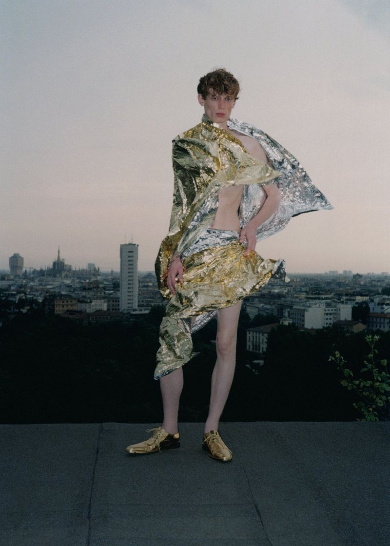 Male fashion model in gold and silver foil outfit poses on rooftop in urban setting with buildings against a pale overcast sky.
