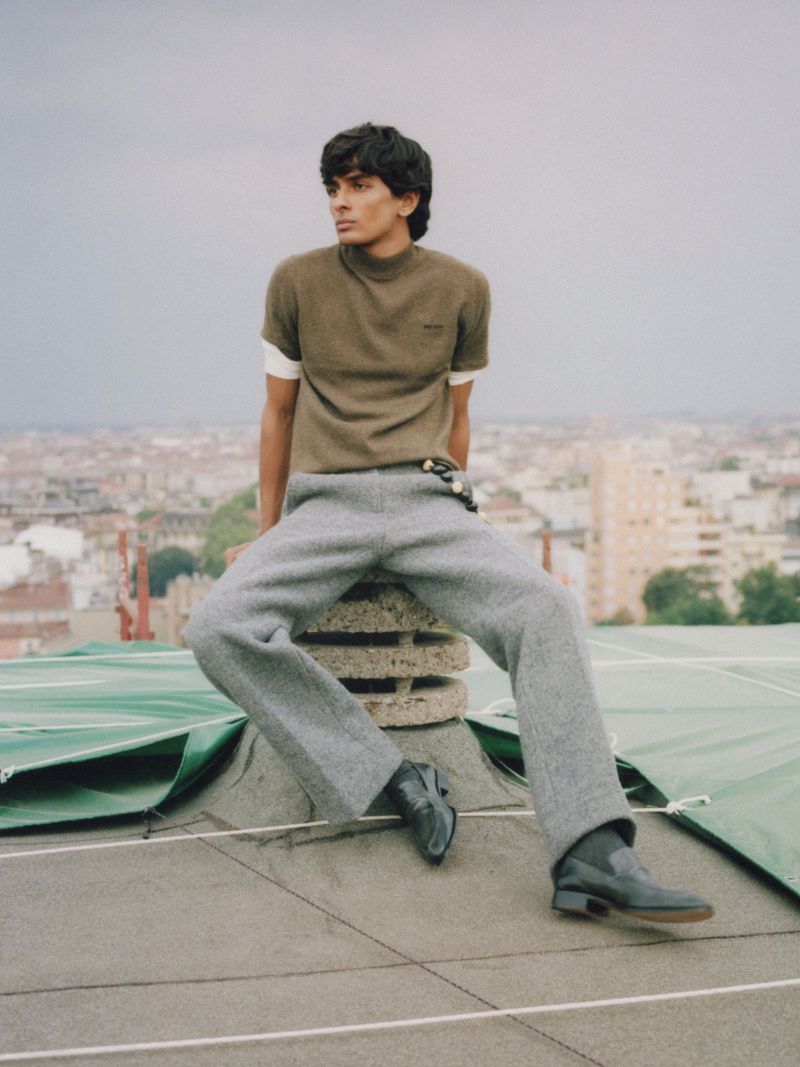 Fashionable young man poses on a rooftop with city views, wearing a casual olive top, gray pants, and black boots in natural light.