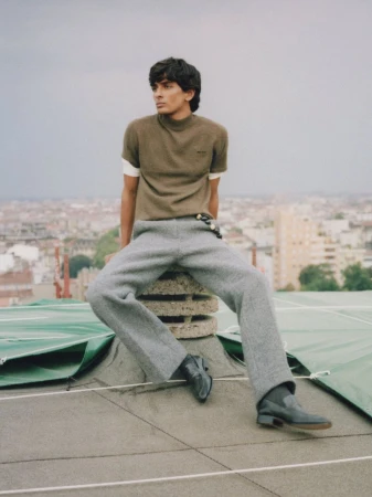 Fashionable young man poses on a rooftop with city views, wearing a casual olive top, gray pants, and black boots in natural light.