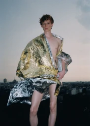 Young male model wrapped in a metallic emergency foil blanket against an overcast city skyline at dusk