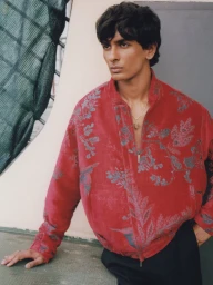 Model wearing a red floral patterned zip-up jacket and black trousers posing outdoors while leaning against a ledge.