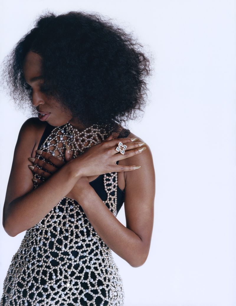 A dark-skinned woman poses in a studio with an open-weave beaded top, a large cocktail ring, and a bright white background for a fashion shot.