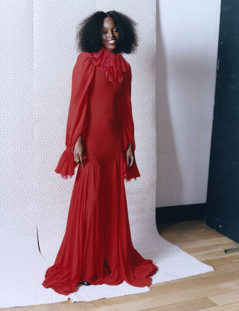 Elegant woman in red gown with frilled collar poses in a studio against polka dot backdrop. The dress has long sleeves and flowing skirt.