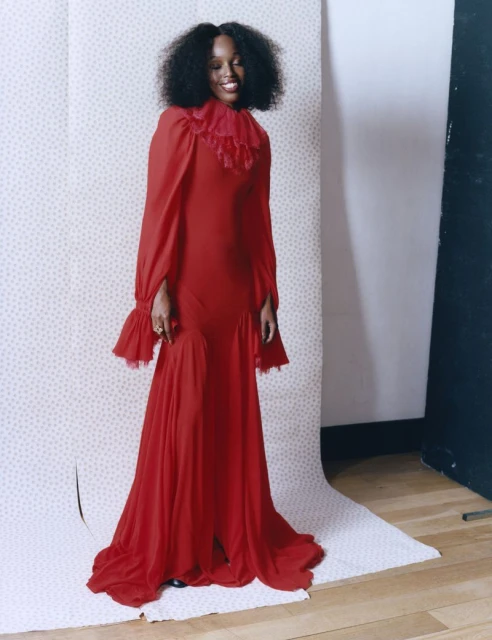 Elegant woman in red gown with frilled collar poses in a studio against polka dot backdrop. The dress has long sleeves and flowing skirt.