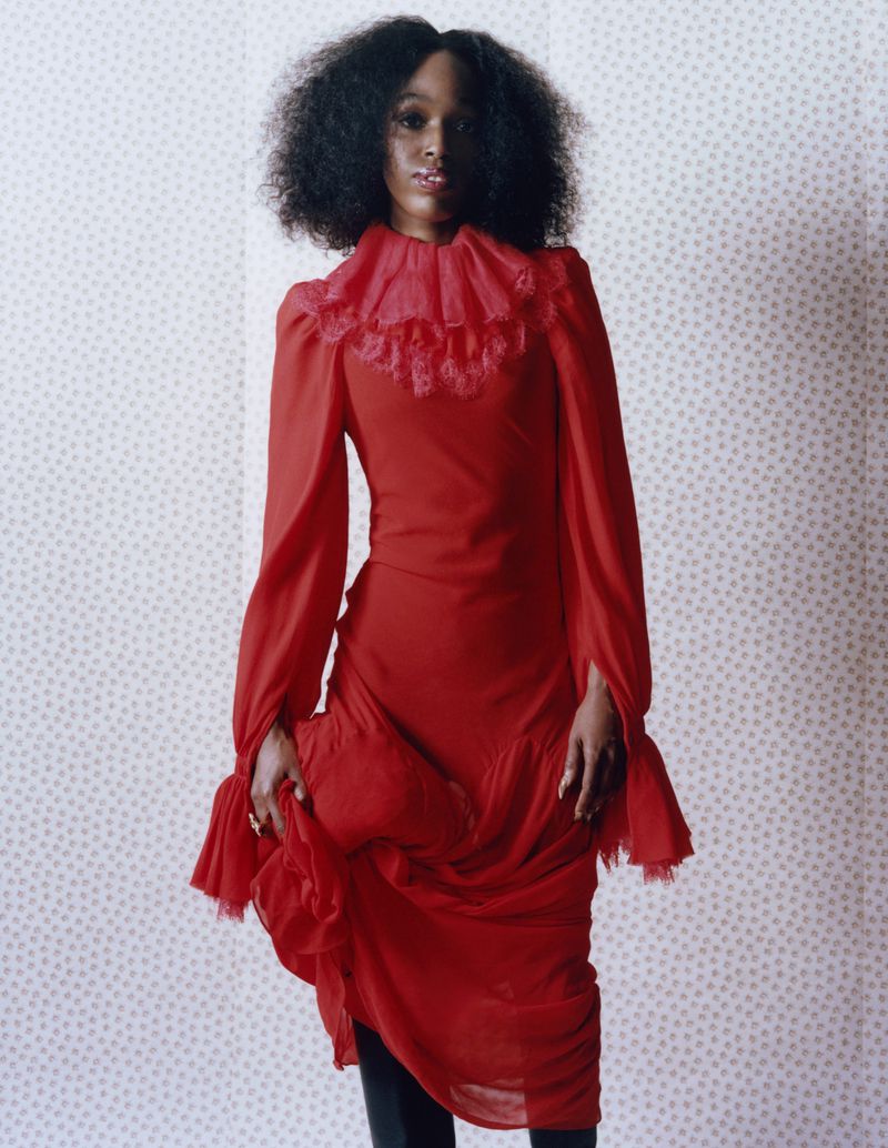A single Black woman stands in a red dress with ruffled collar against a wallpapered background, indoor fashion photography.