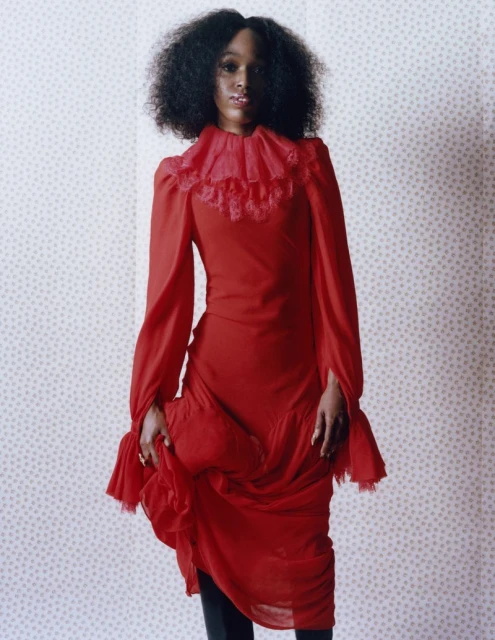 A single Black woman stands in a red dress with ruffled collar against a wallpapered background, indoor fashion photography.