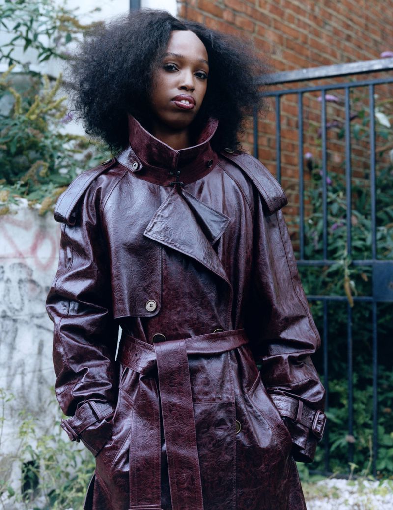 Young adult woman with natural hair wears a belted maroon leather trench coat outdoors in front of a wall and dark green plants.
