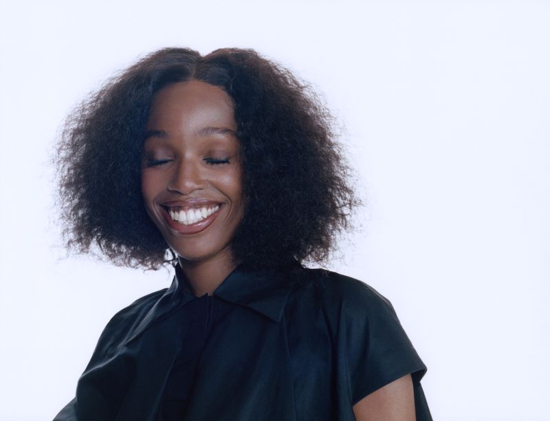 Portrait of a smiling African-American woman with afro hairstyle, wearing a black top, against a plain white backdrop in a studio setting.