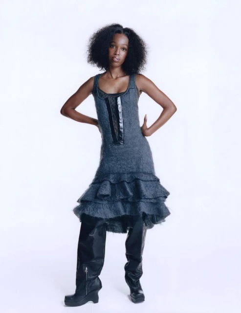 Young woman with dark hair poses in a gray sleeveless dress with black trim, and shiny black thigh-high boots against a white backdrop.