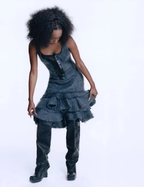 A woman with dark skin and dark curly hair poses in a grey dress and black boots against a white backdrop in a fashion photo.