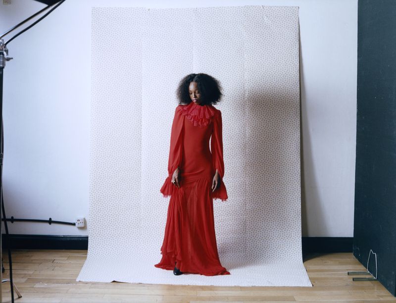 An adult woman with an afro hairstyle poses in a long-sleeved red gown in a photo studio with bright lighting and a patterned backdrop.