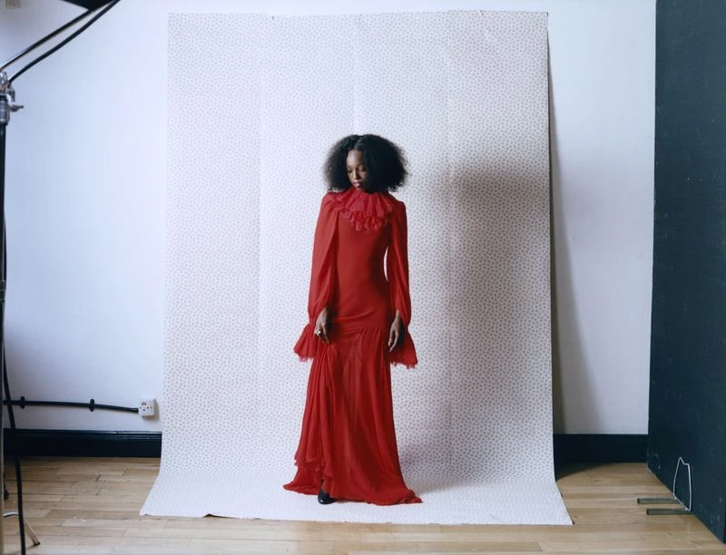 An adult woman with an afro hairstyle poses in a long-sleeved red gown in a photo studio with bright lighting and a patterned backdrop.