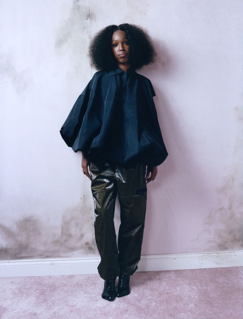 Full-length portrait of a Black female model with afro, wearing an oversized black top and glossy black pants, posing against a textured pink wall.