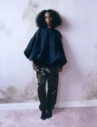 Musician John Glacier posing in a voluminous dark collared top and glossy black trousers against a soft pink mottled wall.