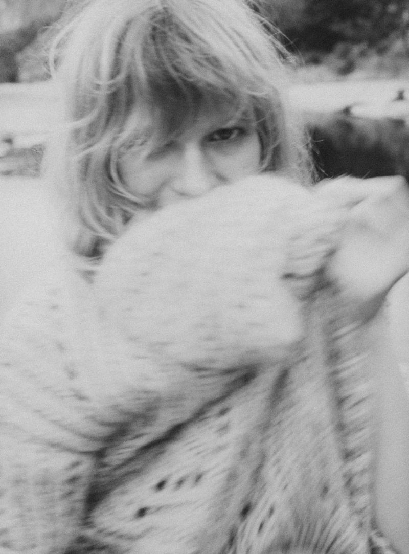 Black and white portrait of a woman with flowing blonde hair partially obscured by a knitted, patterned shawl outdoors.