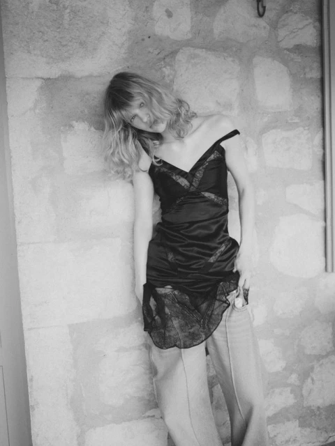 Black and white portrait of a young woman in a black slip dress and pants, leaning against a textured stone wall indoors.