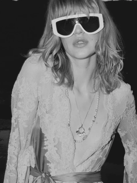 Monochrome portrait of an adult female wearing wide white sunglasses, long necklace, and a lace-trimmed robe or dress, posing indoors.