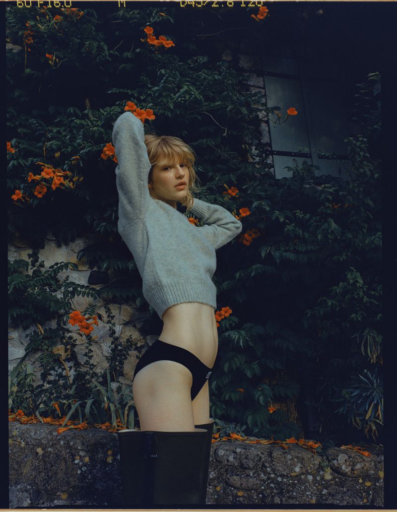 Blonde model in a bucket posing against a stone wall with orange flowering vines, wearing a grey sweater and black bottoms, outdoor setting.