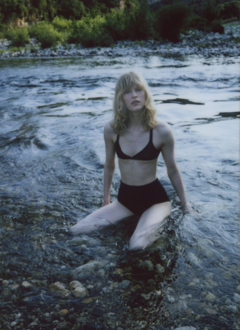 Young woman with blonde hair sits in a bikini in a shallow river with rocky bottom and a forest in the background on an overcast day.