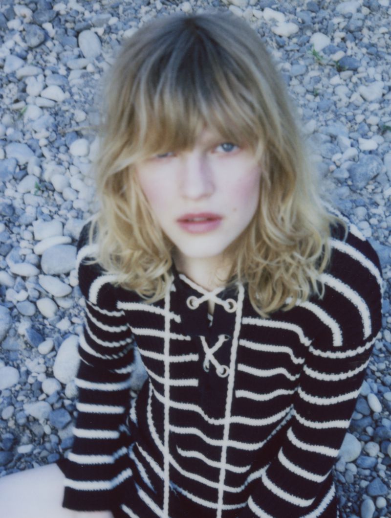 Blonde female fashion model in striped sweater poses outside on a pebble beach in medium shot, looking directly at camera.