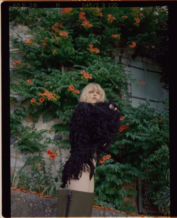 Woman with shaggy black coat poses in front of a green vine-covered wall with orange trumpet flowers in an outdoor setting.